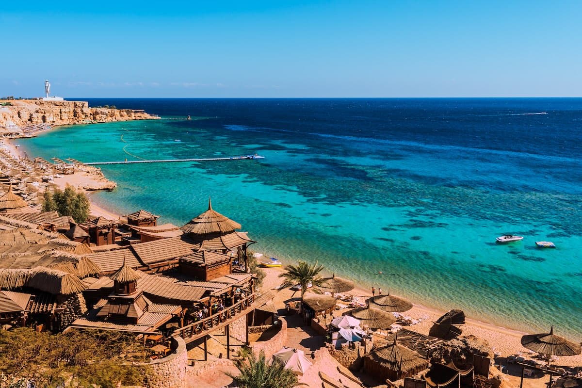 Aerial view of a bay near Sharm el-Sheikh on the Red Sea — thatched beach restaurants on stilts, turquoise lagoon water, jetties and offshore reefs.